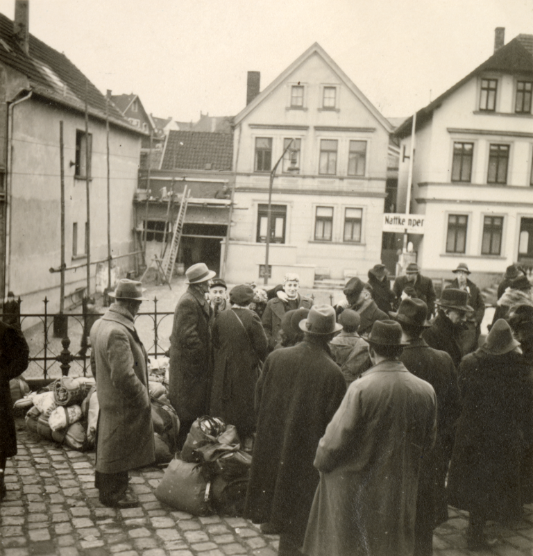 Die am 13. Dezember 1941 deportierten Jüdinnen und Juden warten am Bahnhof, in Sichtreichweite einiger Wohnhäuser.