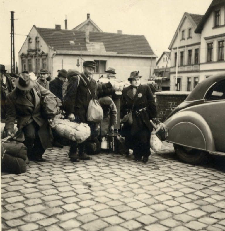 Das Original zeigt eine wartende Gruppe in Nähe des „Kyffhäusers“ von vorn, von der allerdings niemand zum Fotografen blickt; rechts am Bildrand halb zu sehen ist ein Auto mit Schiebeverdeck, das am Ende des dritten Kriegsjahres zweifelfrei den zuständigen Gestapofunktionären zuzuordnen ist.