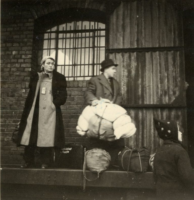 Vor dem vergitterten Fenster des Zoll- und Güterschuppens wartet das Ehepaar Jenny und Heinrich Mayer auf den Deportationszug. Laut Aufschrift enthält der Koffer zwischen ihnen Werkzeug und Töpfe. Der Mann hält eine riesige Tasche.
