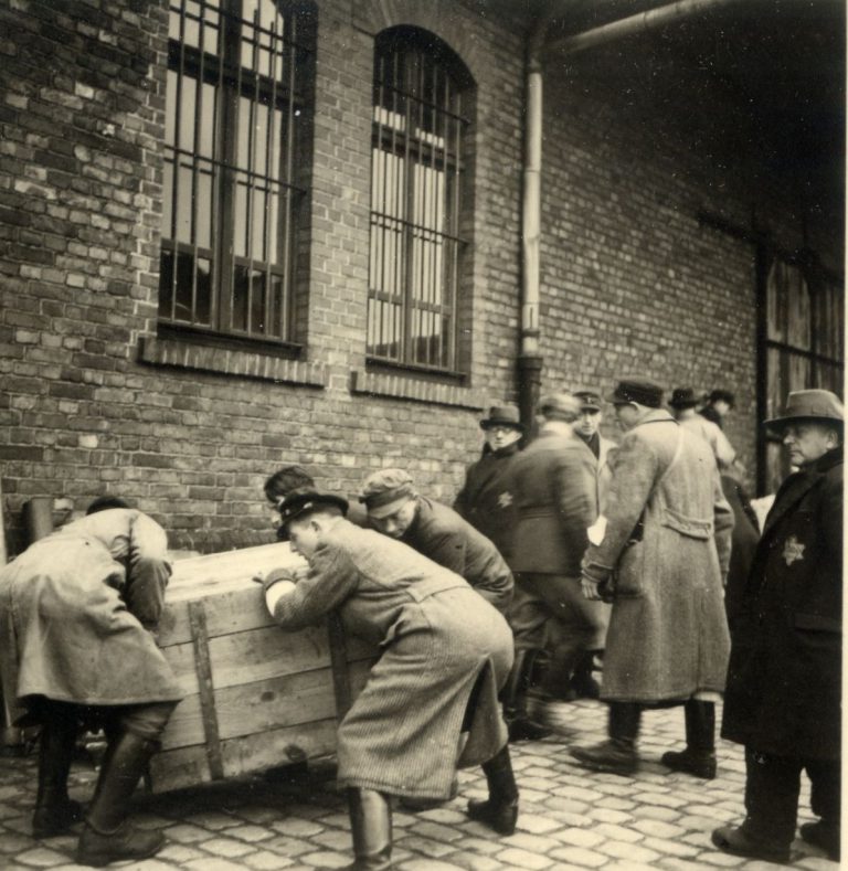 Eine Gruppe jüdischer Männer, die sich um eine Kiste mit Schwergepäck mühen. In der Bildmitte ist Bielefelder Riga-Überlebende Artur Sachs zu erkennen. Am linken Arm trug er eine weiße Binde, die als Mitglied des jüdischen Ordnungsdienstes während der Fahrt des Deportationszuges kennzeichnete.
